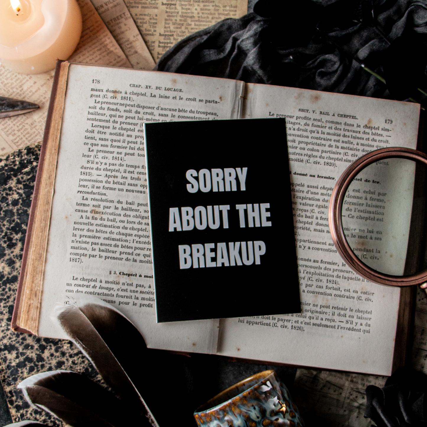 A greeting card with 'SORRY ABOUT THE BREAKUP' written in white text on a black background, placed on an open book surrounded by a candle, a feather, and an envelope, with a mirror reflecting a candle.
