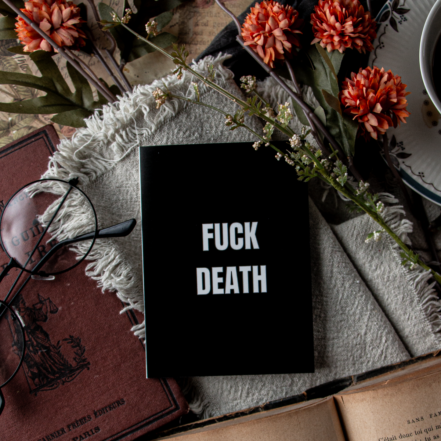 Black book with 'FUCK DEATH' text on a table with flowers and books.