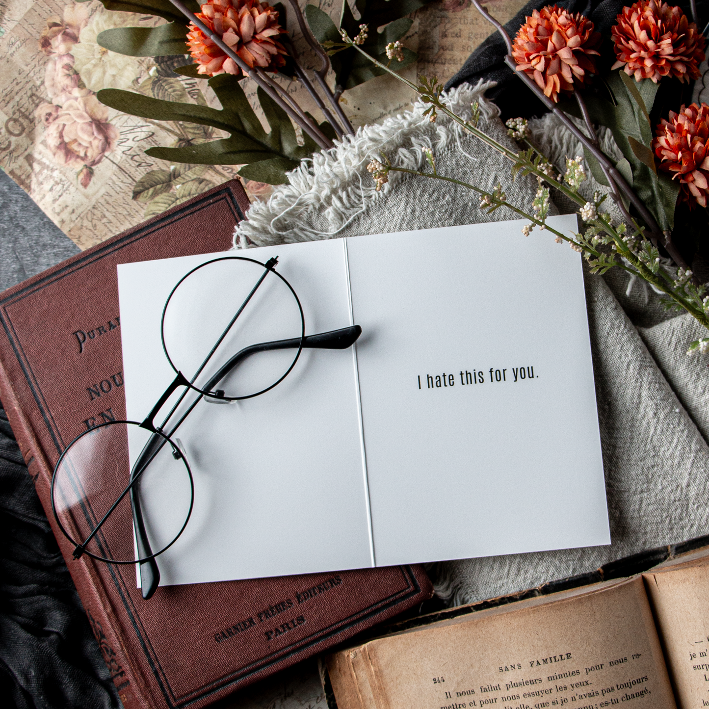 Open book with a card reading 'I hate this for you' on a surface with flowers and glasses.