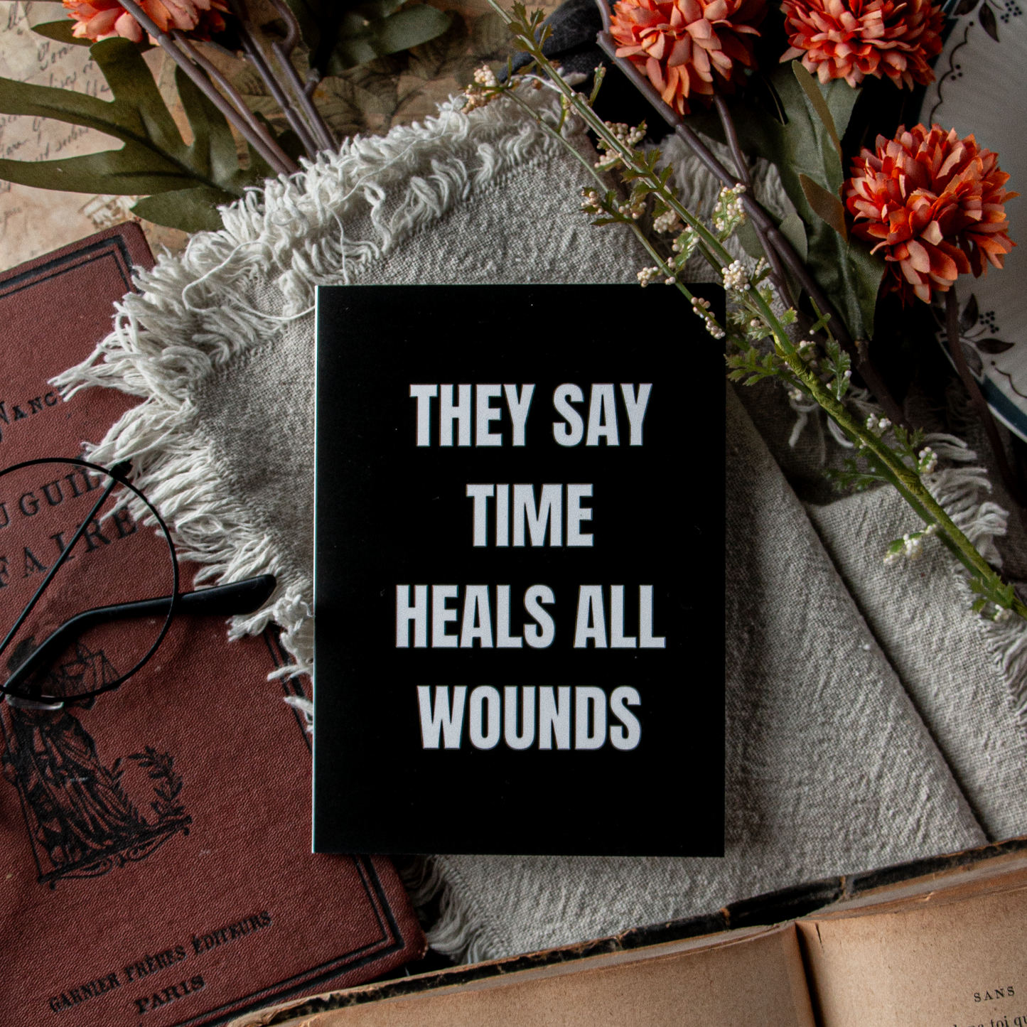 Black sign with 'THEY SAY TIME HEALS ALL WOUNDS' text on a textured surface with flowers and books.