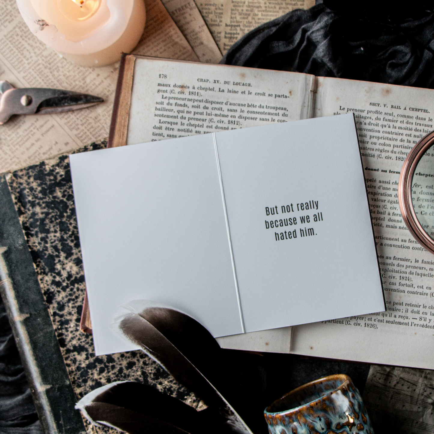 A humorous breakup card with the text 'But not really, we all hated him' placed inside an open book, surrounded by a cup, feathers, and a candle.