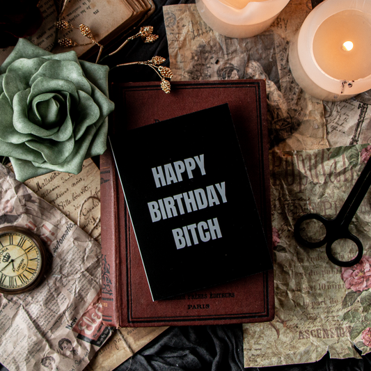 A greeting card with 'HAPPY BIRTHDAY BITCH' written in white text on a black background, displayed on a table with candles, a clock, and other decorations.