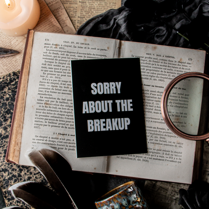 A greeting card with 'SORRY ABOUT THE BREAKUP' written in white text on a black background, placed on an open book surrounded by a candle, a feather, and an envelope, with a mirror reflecting a candle.