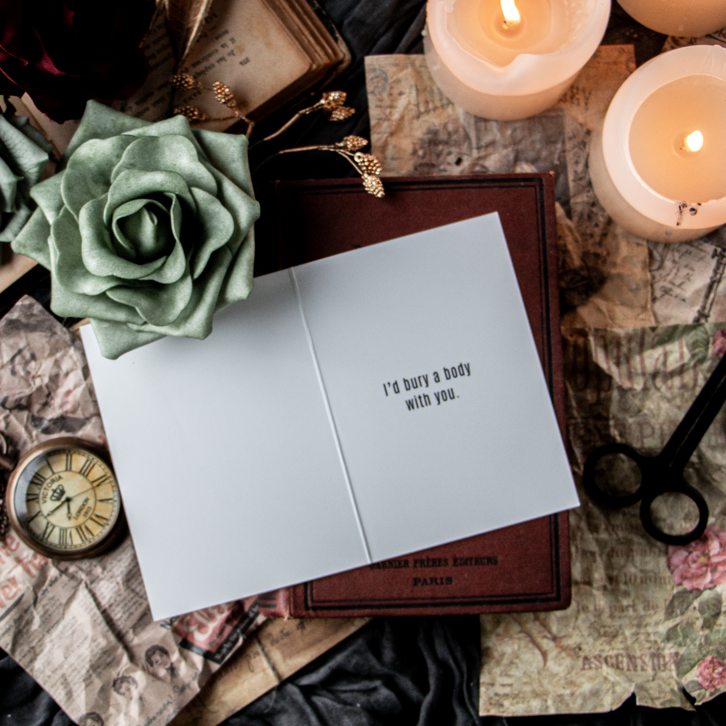 An overhead shot of a birthday card with the text 'HAPPY BIRTHDAY BITCH' on the front and 'I’d bury a body with you' on the inside, placed on a surface with candles, a flower, and vintage-style props.