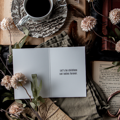 A greeting card with the text 'Let's be childless cat ladies' opened to a person, placed on a table with a cup of coffee, books, and decorative flowers.