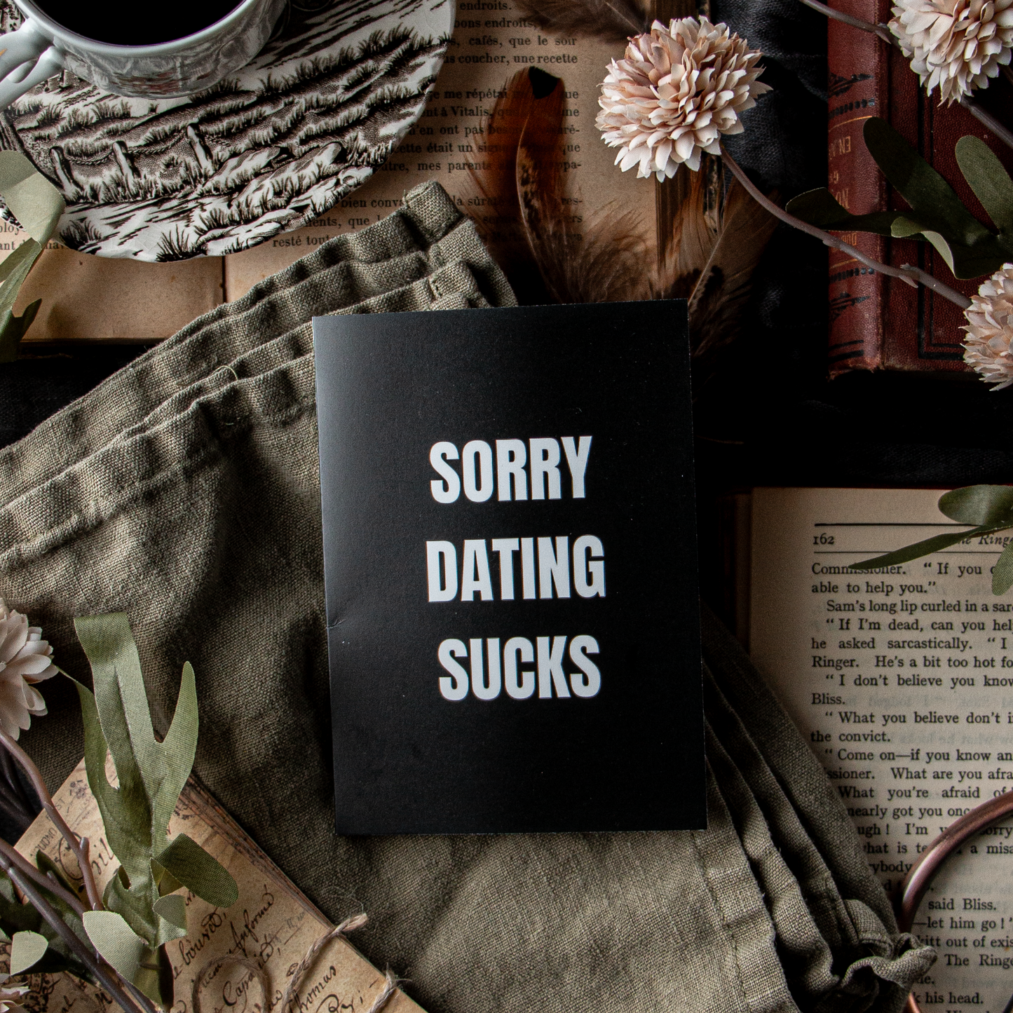 A greeting card with the text 'SORRY DATING SUCKS' placed on a table with a cup of coffee, flowers, and vintage books.