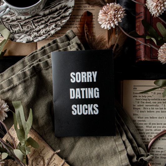 A greeting card with the text 'SORRY DATING SUCKS' placed on a table with a cup of coffee, flowers, and vintage books.