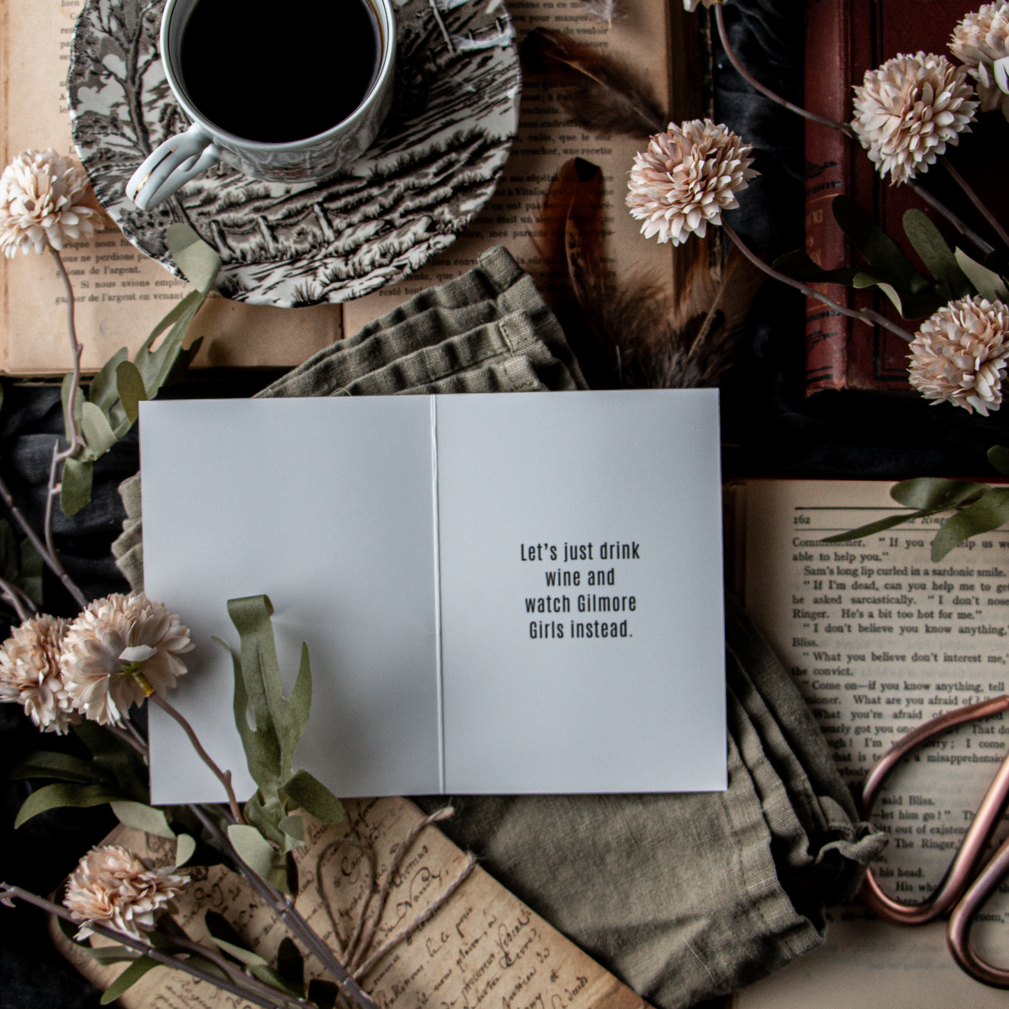 A greeting card with the text 'Let's drink wine' opened to reveal a blank interior, placed on a surface with a cup of coffee, books, and decorative flowers.