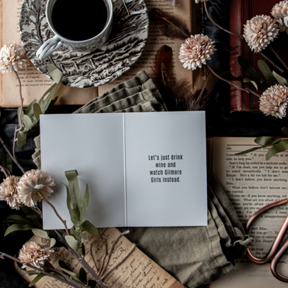 A greeting card with the text 'Let's drink wine' opened to reveal a blank interior, placed on a surface with a cup of coffee, books, and decorative flowers.