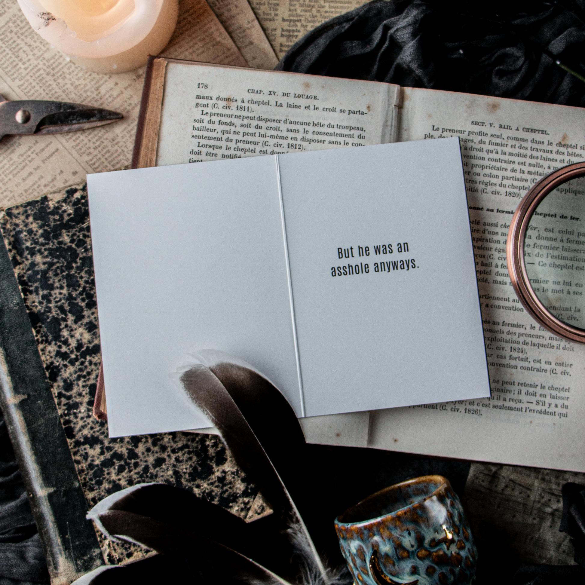 A greeting card with the text 'SORRY ABOUT THE BREAKUP' displayed on the front, placed on a table alongside a candle, a book, and a feather pen.