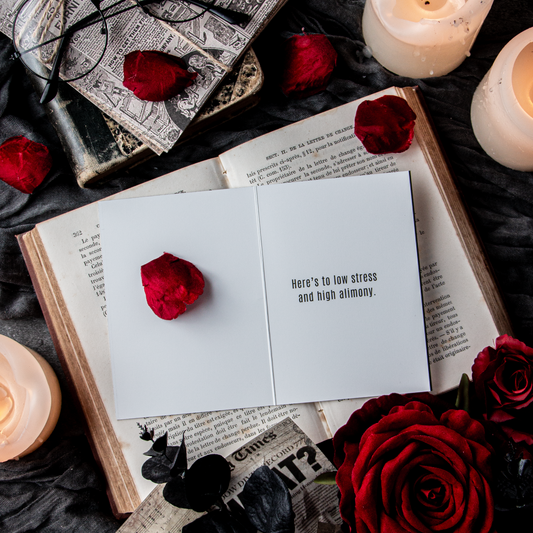 A greeting card with the text 'CONGRATS ON YOUR FREEDOM! HERE'S TO LOW STRESS AND HIGH ALIMONY.' placed on a table with a book, candles, and rose petals around it.