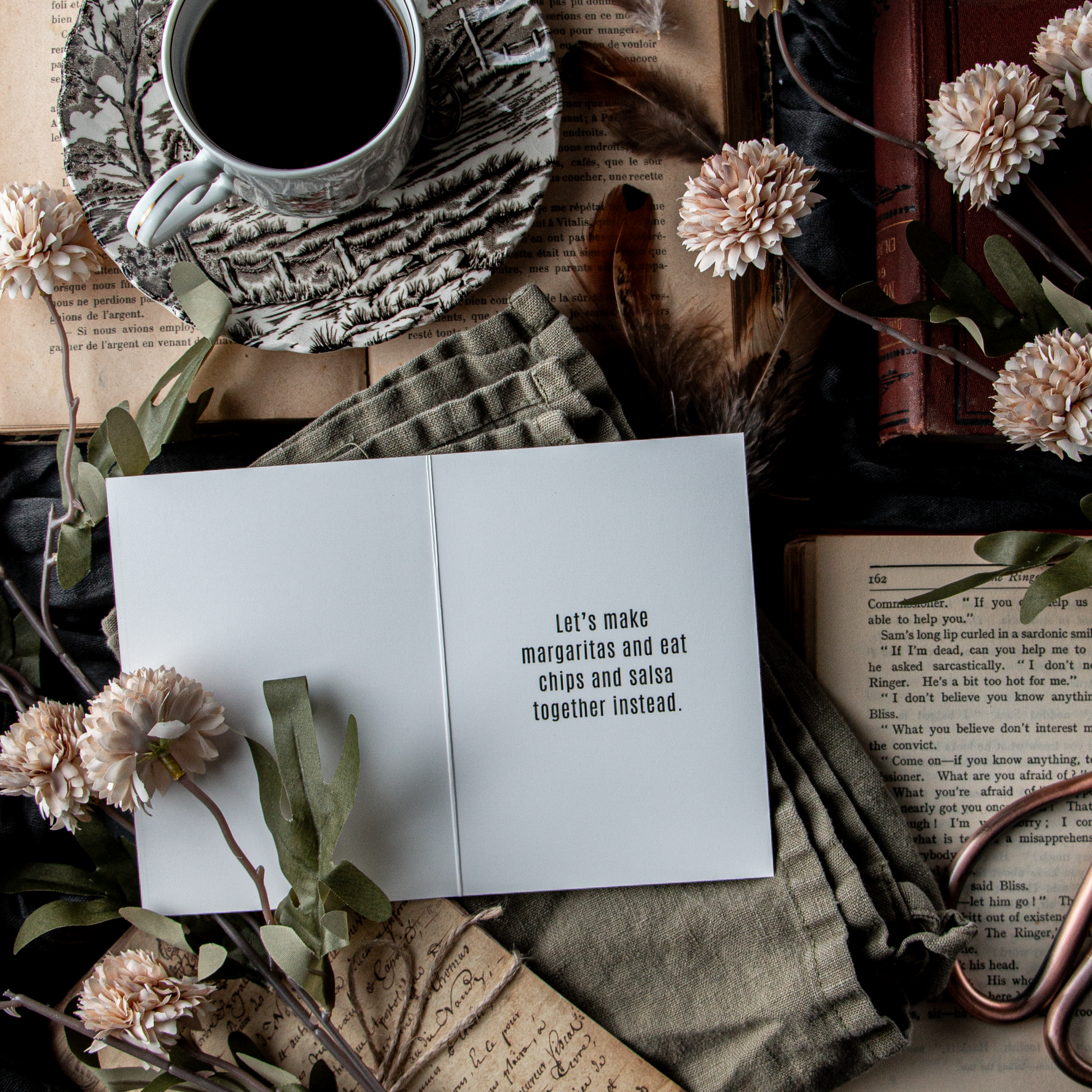 A greeting card with the text 'SORRY DATING SUCKS' placed on a table with a cup of coffee, books, and decorative flowers.