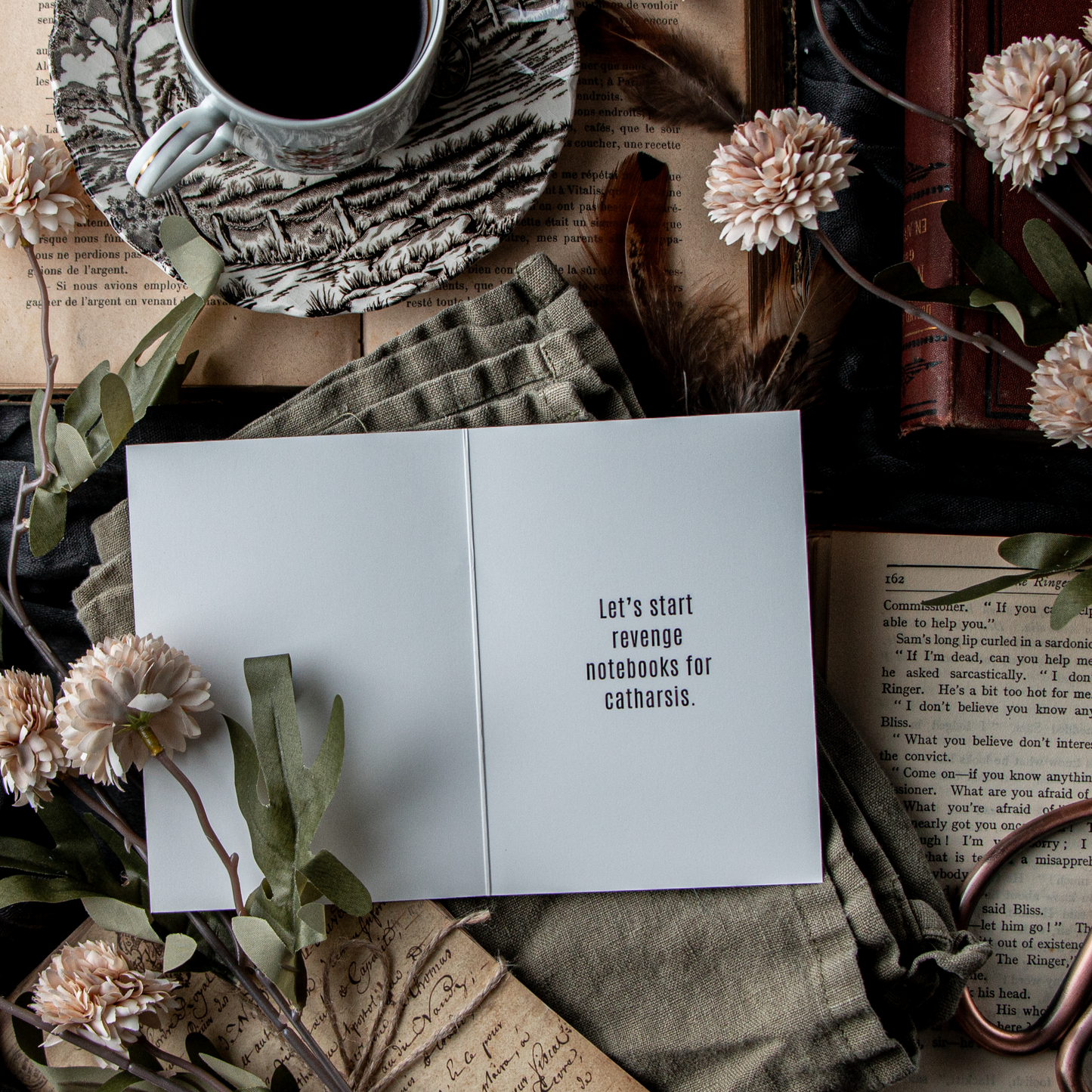 A greeting card with the text 'Let's start revenge notebooks for catharsis' displayed open, surrounded by a cup of coffee, books, and decorative flowers laid out on a table.