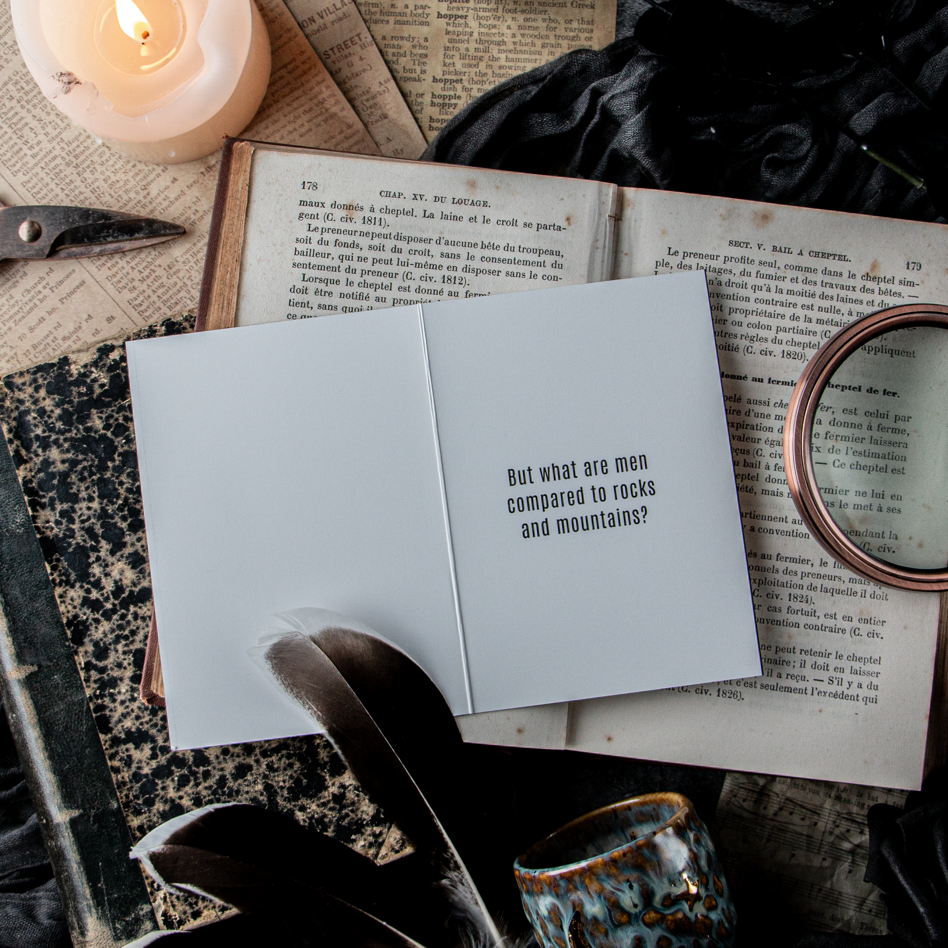 A greeting card laid out on a flat surface with a candle and books, partially opened, displaying a humorous message suited for a breakup, surrounded by a feather and a small decorative bell.