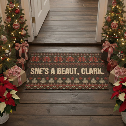 Decorative doormat with 'She's a Beaut, Clark' text on a wooden floor with Christmas trees and poinsettias.