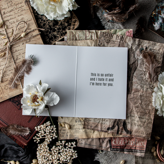 A greeting card with 'F*ck Cancer' written in bold white letters on the front, placed on a table with a floral pattern alongside other decorative items.